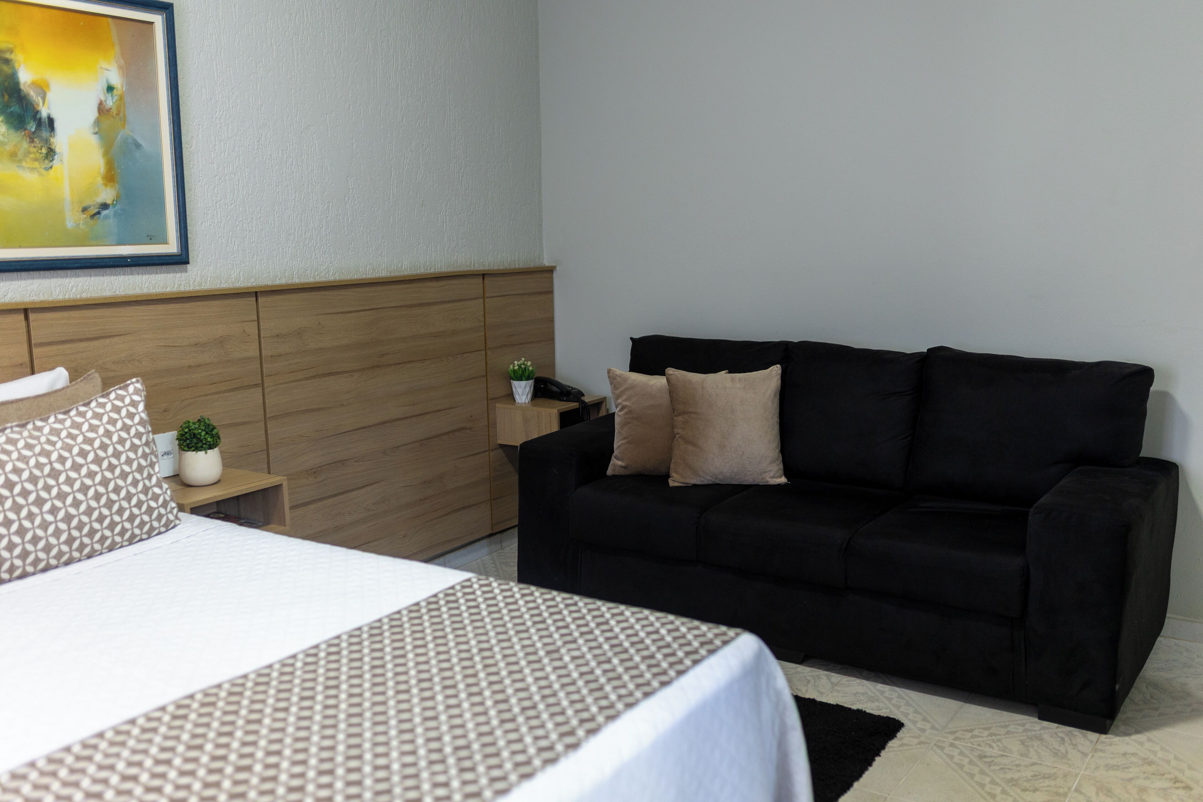 Ein Hotelzimmer mit einem Bett, einer schwarzen Couch, einem Holz-Kopfteil, einer kleinen Pflanze und einem Gemälde an der Wand.