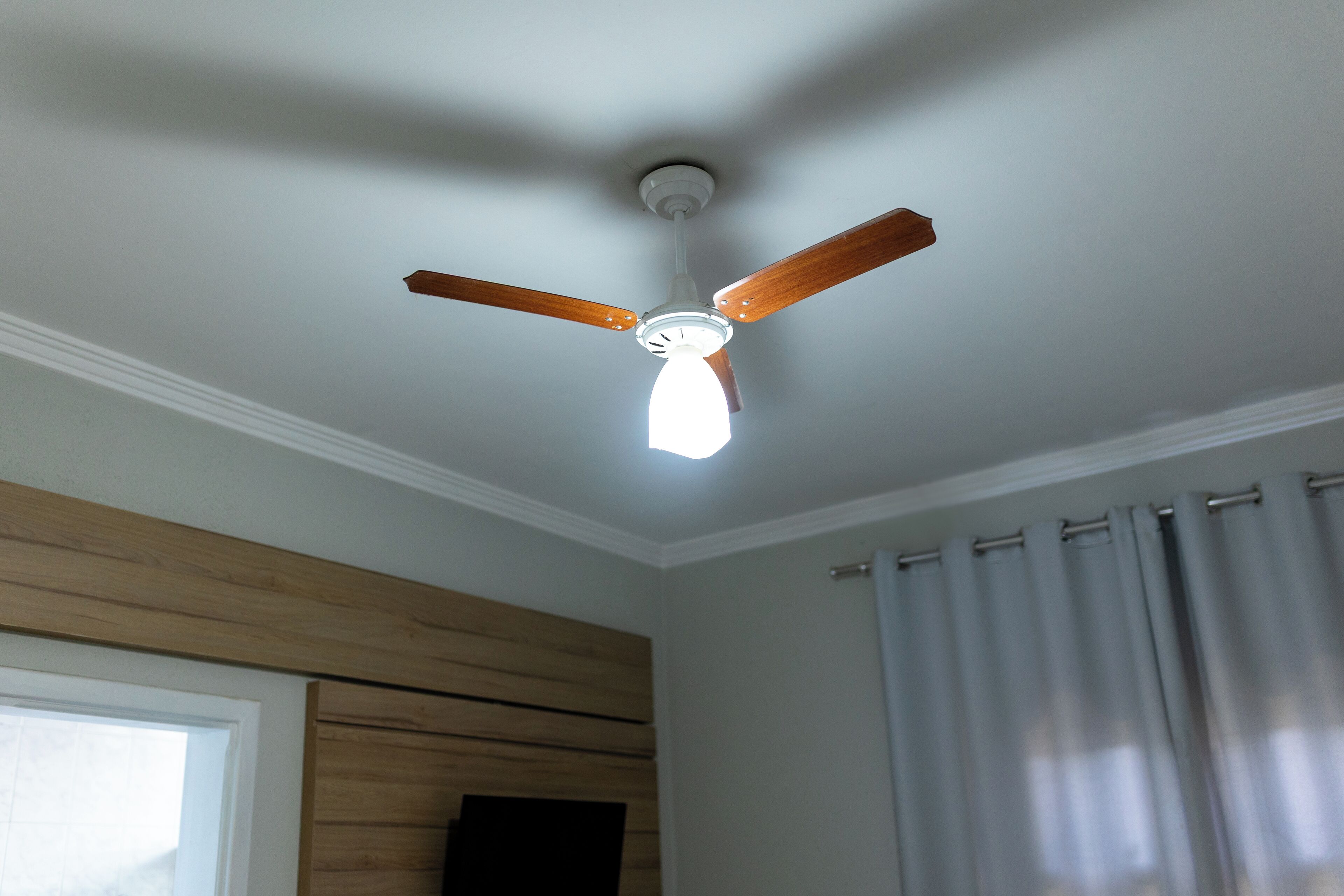 Un ventilador de techo con aspas de madera y una lámpara en una habitación con paneles de madera y una ventana con cortinas.