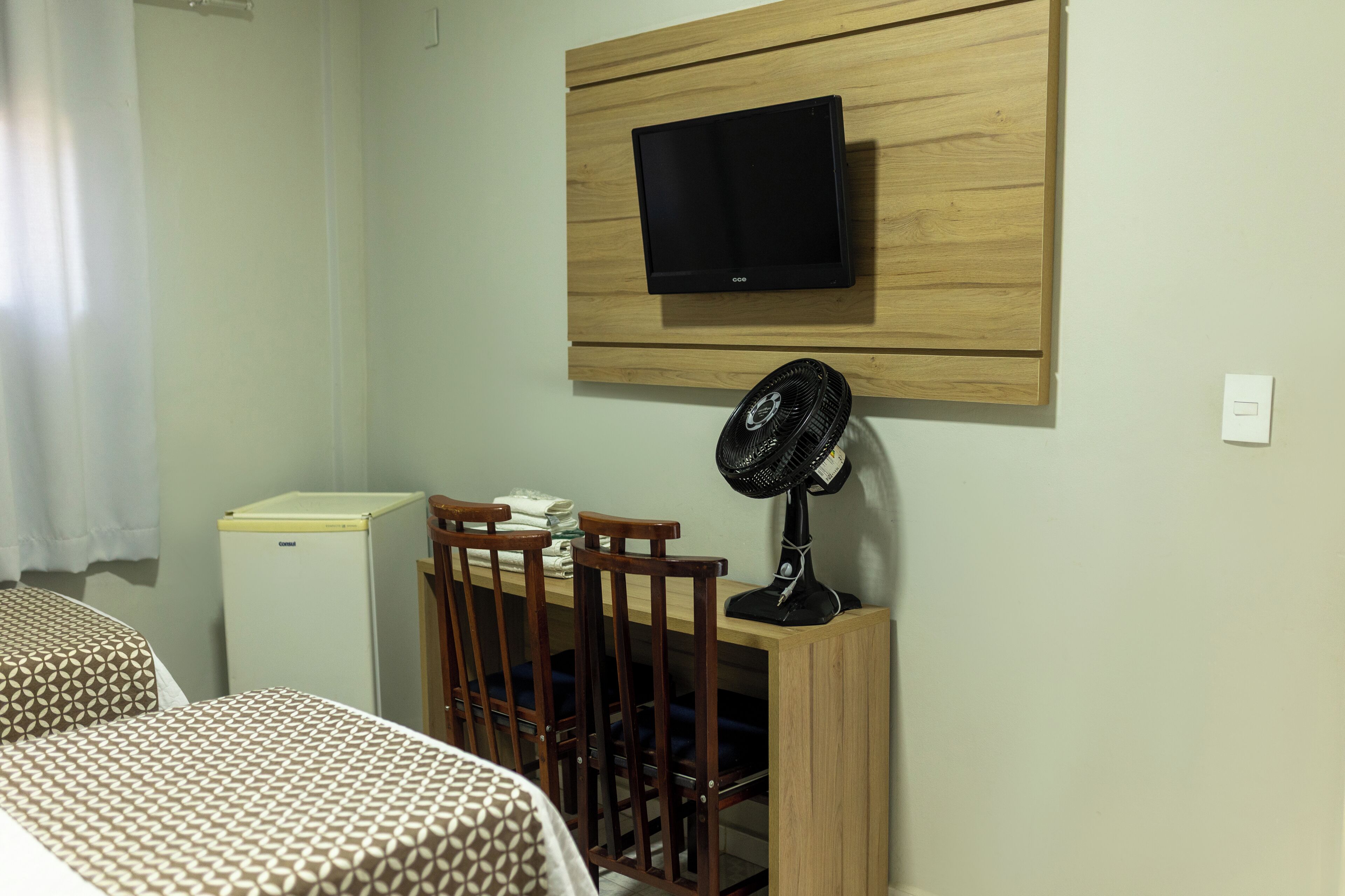 Ein Zimmer mit Bett, Schreibtisch mit Stuhl, kleinem Kühlschrank, Wand-Fernseher und Standventilator.