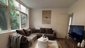 Un salon moderne avec un canapé d’angle, une table basse ronde, un téléviseur à écran plat et de grandes fenêtres donnant sur un paysage verdoyant.