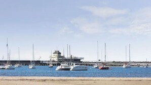 Un port de plaisance où sont amarrés plusieurs voiliers, une jetée qui s’avance dans l’eau et, au fond, un bâtiment surmonté d’une tour.