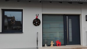 Une maison moderne avec une porte d’entrée sombre, le numéro « 62 » au-dessus de la porte, une guirlande accrochée au mur et une boîte aux lettres.