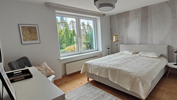 Une chambre à coucher avec un grand lit, un bureau, une chaise, un radiateur et une fenêtre donnant sur des arbres.