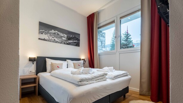 Un lit bien fait, avec du linge de lit blanc, une table de chevet avec une lampe, un tableau accroché au mur et une fenêtre ornée de rideaux rouges.