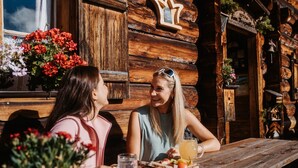 Twee personen zitten aan een houten tafel met eten en drinken, voor een eenvoudig houten gebouw met een bord.