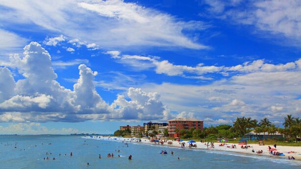 Una spiaggia con persone che nuotano e prendono il sole, edifici sullo sfondo e un cielo parzialmente nuvoloso.