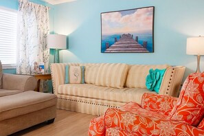 A living room with a striped sofa, a red patterned chair, a painting of a pier, and a lamp on a side table.