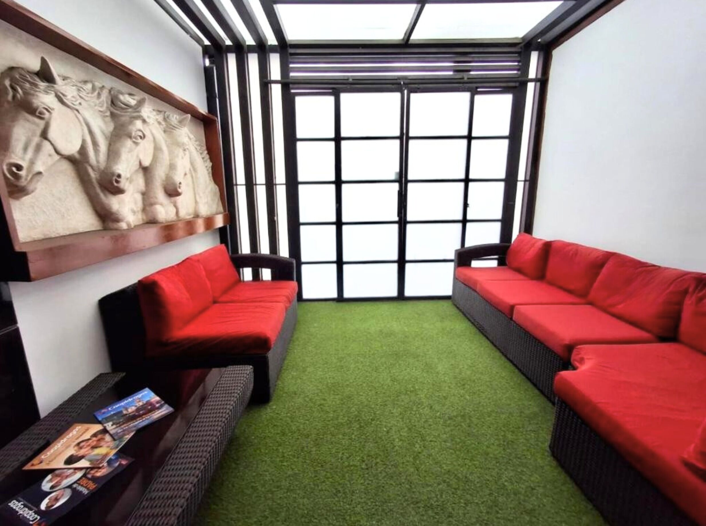 Um quarto com dois sofás vermelhos, piso de grama sintética verde, uma escultura de cavalo na parede e uma grande janela com molduras pretas.