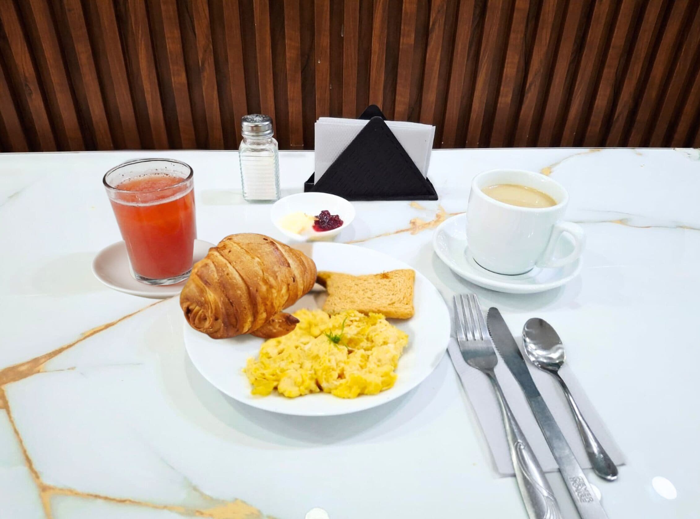 A breakfast setting with scrambled eggs, toast, a croissant, a glass of juice, a cup of coffee, and a salt shaker on a marble table.