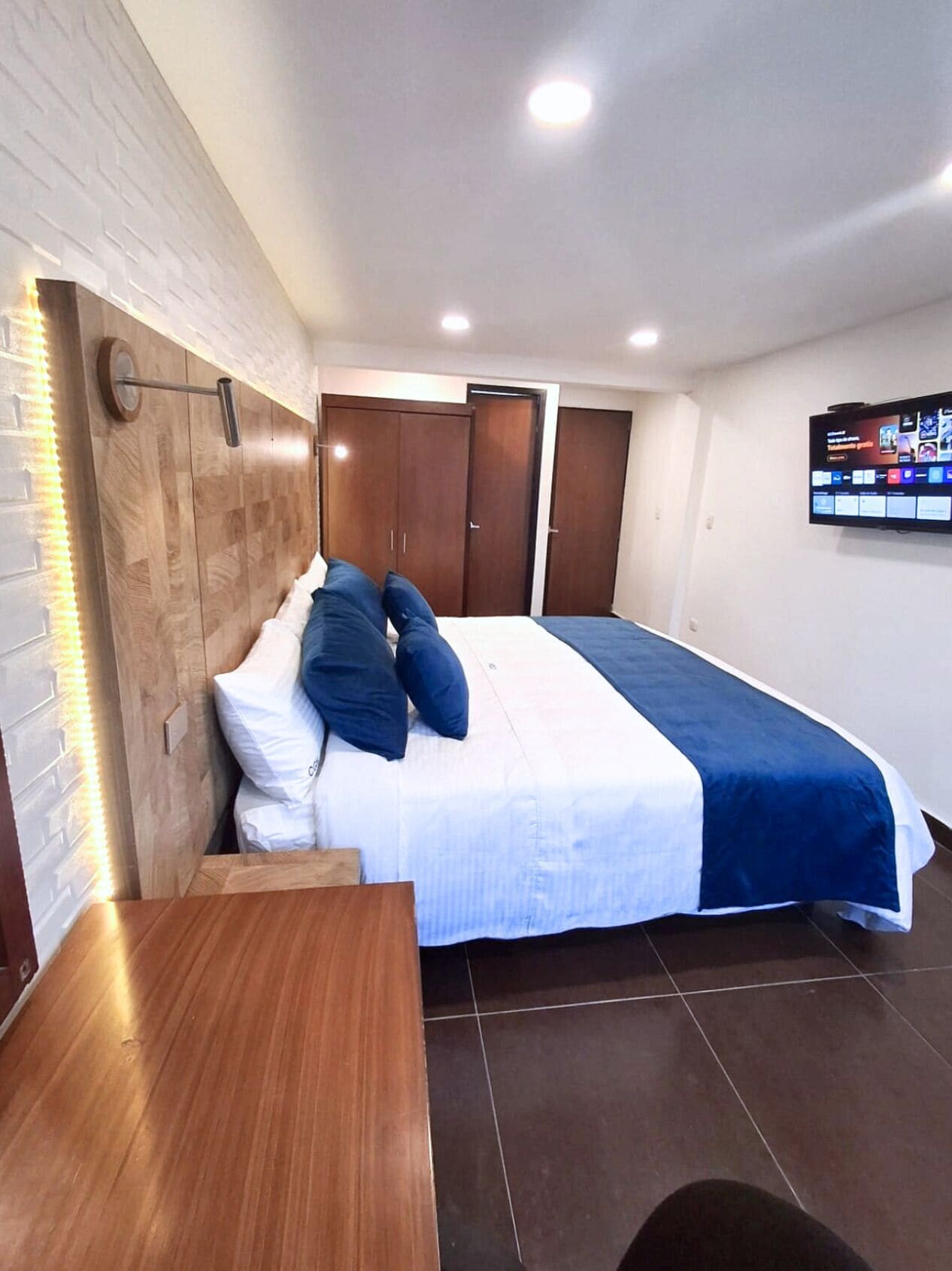 A hotel room with a bed, a wooden headboard, a TV mounted on the wall, and a wooden table.