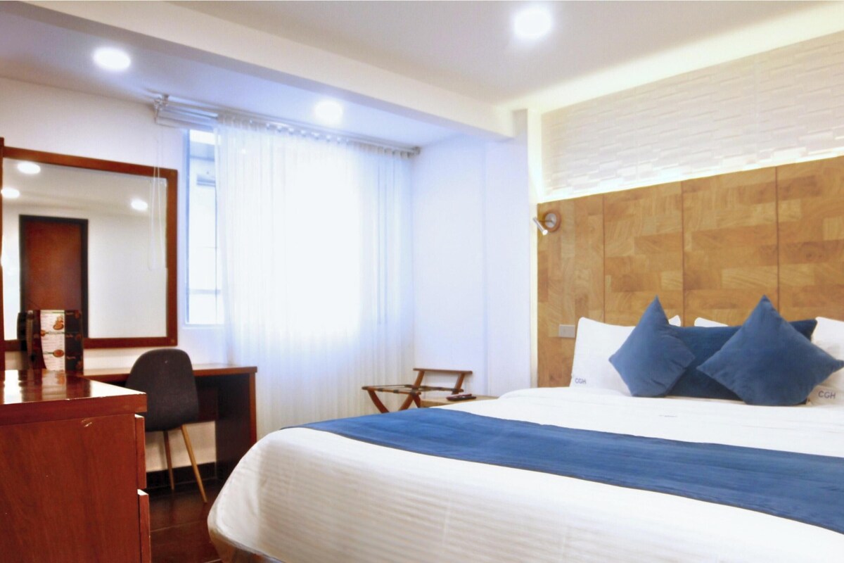 A hotel room with a bed, a desk, a chair, a mirror, and a window with curtains.