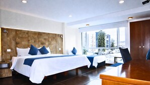 A hotel room with a large bed, a desk, and a view of buildings through the window. - Hotel CGH Travel INN Aeropuerto (Bogota)
