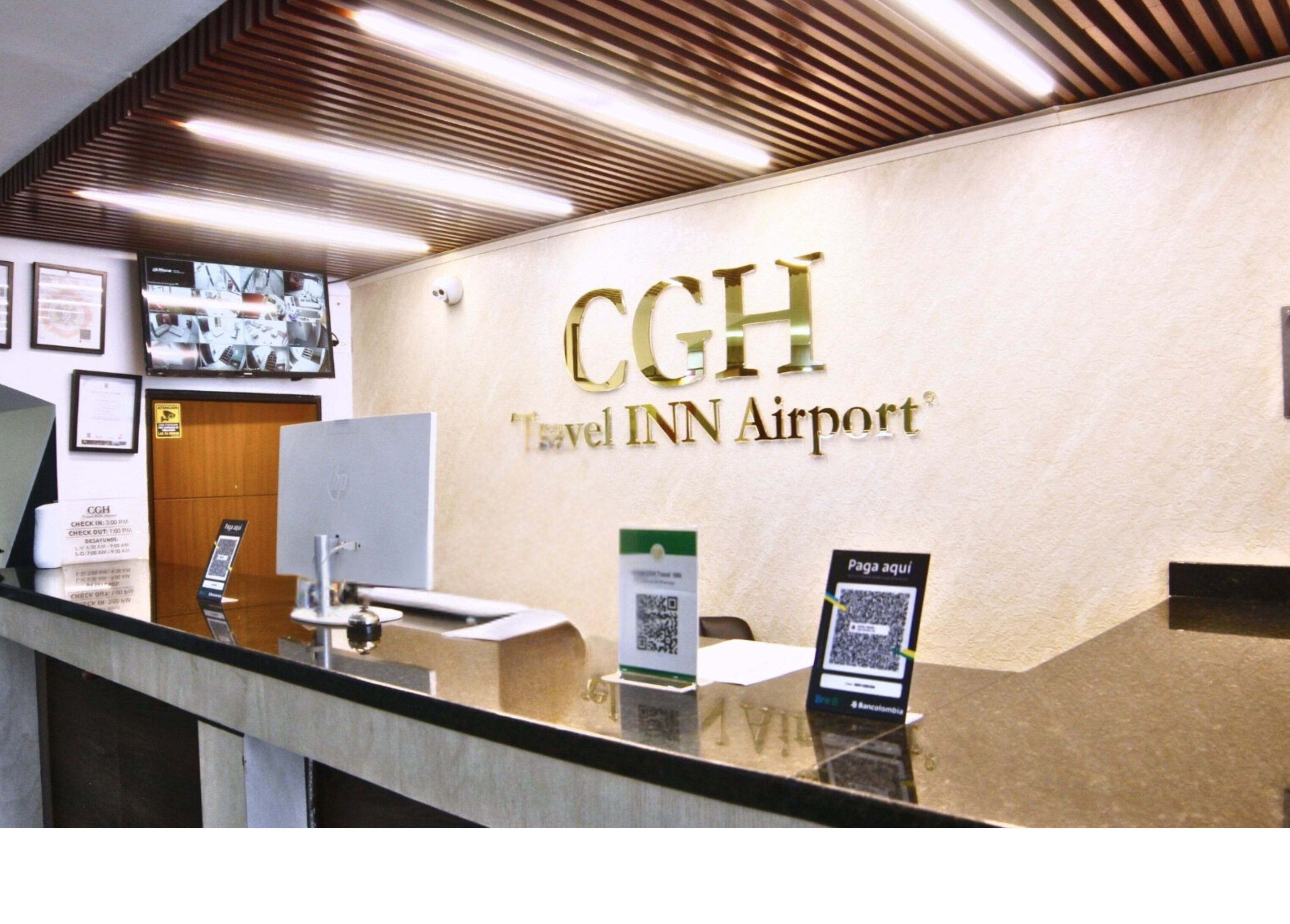 Un mostrador de recepción con un cartel que dice 'CGH Travel Inn Airport', una computadora, una foto enmarcada y un código QR que indica 'Paga aquí'.
