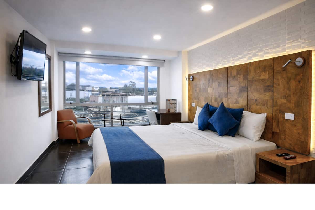 A modern hotel room with a large bed, a TV mounted on the wall, a wooden headboard, and a view of a cityscape through large windows.