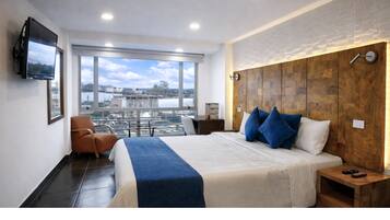 A modern hotel room with a large bed, a TV mounted on the wall, a wooden headboard, and a view of a cityscape through large windows.