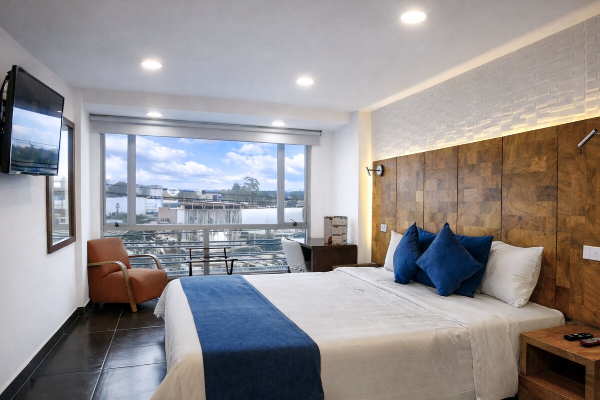 A modern hotel room with a large bed, a TV mounted on the wall, a wooden headboard, and a view of a cityscape through large windows.