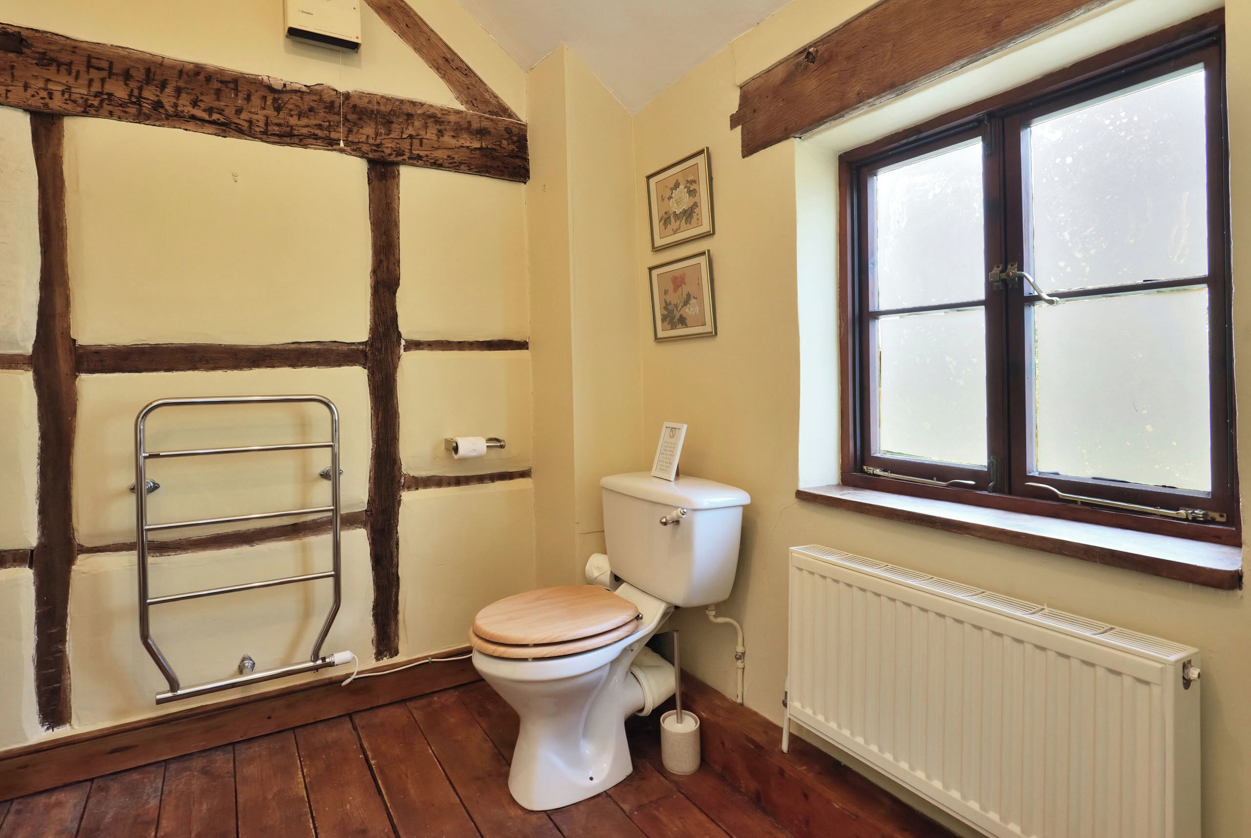 Un bagno con pavimento in legno, un WC bianco con sedile in legno, un termosifone e una finestra con vetro smerigliato.