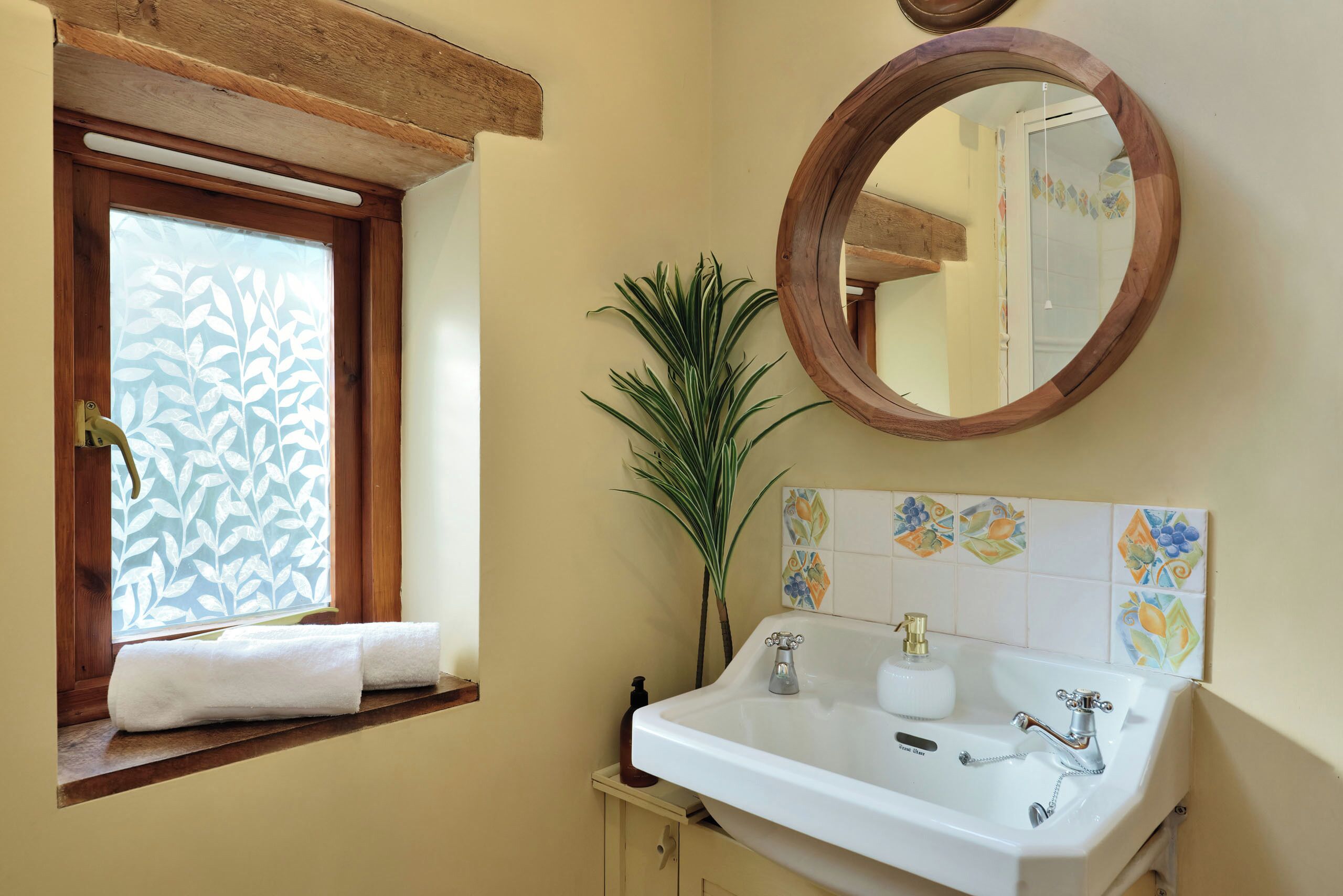 Une salle de bain avec une fenêtre en bois, un lavabo à deux robinets, un miroir et une plante en pot.