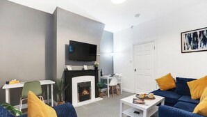 A modern living room with a fireplace, a flat-screen TV mounted on the wall, a dining area with a white table and chairs, and a blue sofa with yellow cushions. - Cosy 2-Bedroom Apartment, comfortably sleeps 5 (Gateshead)