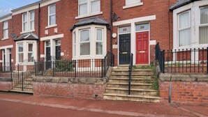 A row of red brick terraced houses with white-framed windows, numbered doors, and black metal railings. - Cosy 2-Bedroom Apartment, comfortably sleeps 5 (Gateshead)