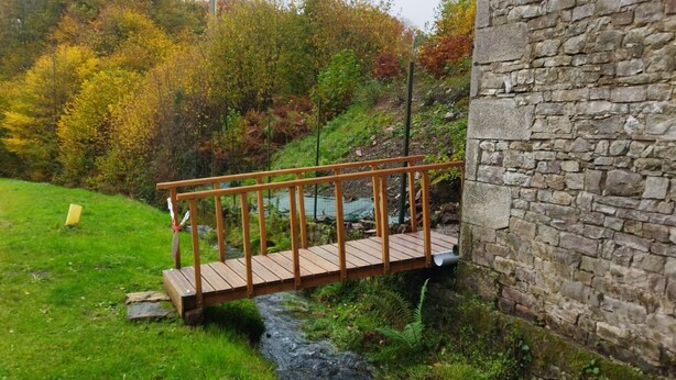 Un puente de madera sobre un arroyo, un muro de piedra y un sendero que se adentra en un área boscosa.