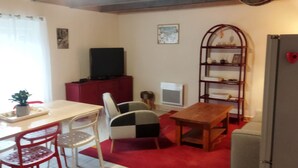 Un espacio habitable compacto con una mesa de comedor, sillas, un televisor pequeño en un mueble, una estantería y un refrigerador.