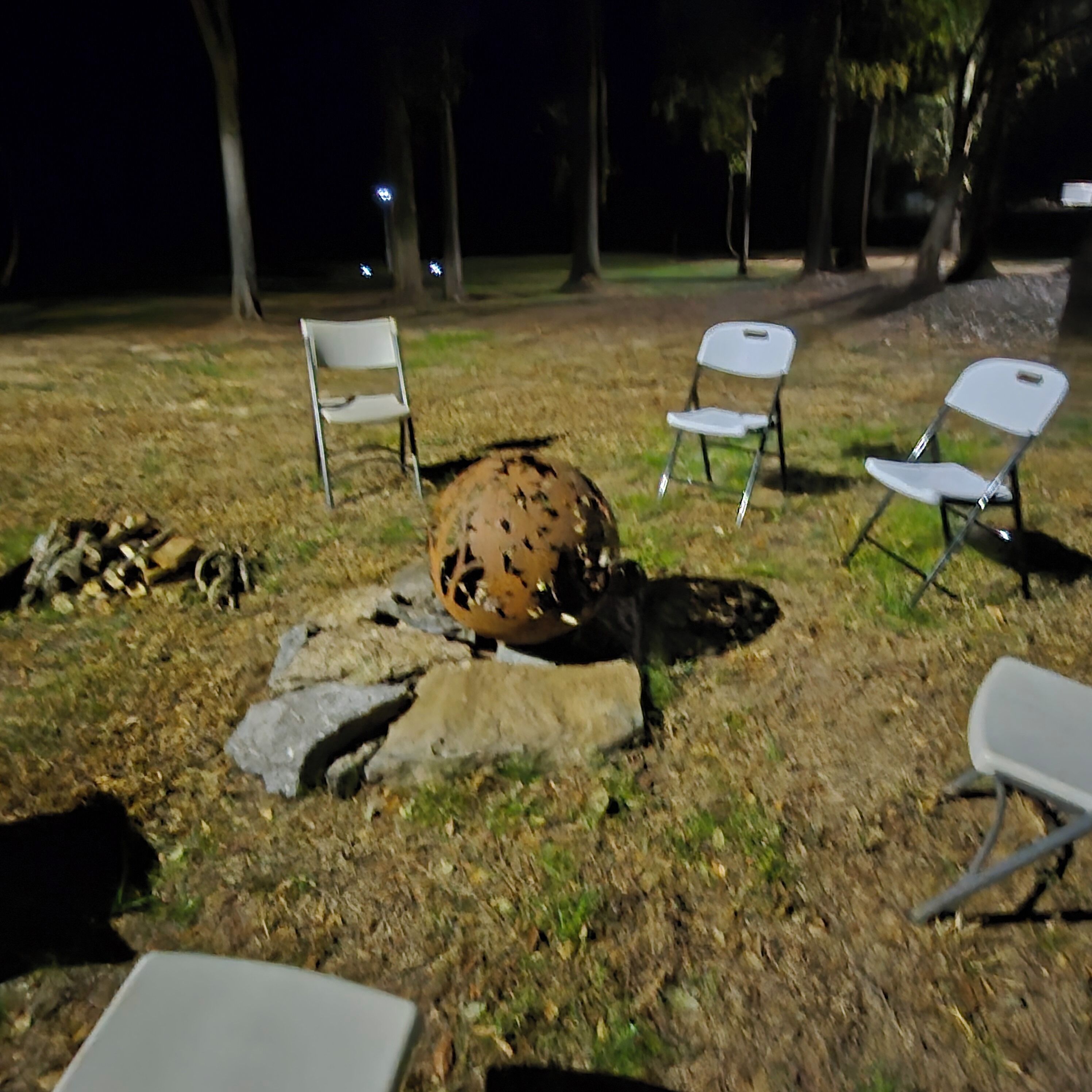 A campsite with a fire pit surrounded by rocks and logs, and four white folding chairs arranged around it.
