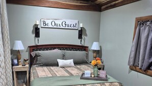 A cozy bedroom with a bed, two bedside tables, a wall-mounted sign, and a window with curtains. - Owl's Nest, a rustic tiny house located 3.5 miles off I-65 in Evergreen, Alabama (Evergreen)
