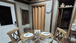 A dining area with a round glass table surrounded by four chairs, each with a woven seat. A wooden door with a window and a wall-mounted clock are visible. - Owl's Nest, a rustic tiny house located 3.5 miles off I-65 in Evergreen, Alabama (Evergreen)