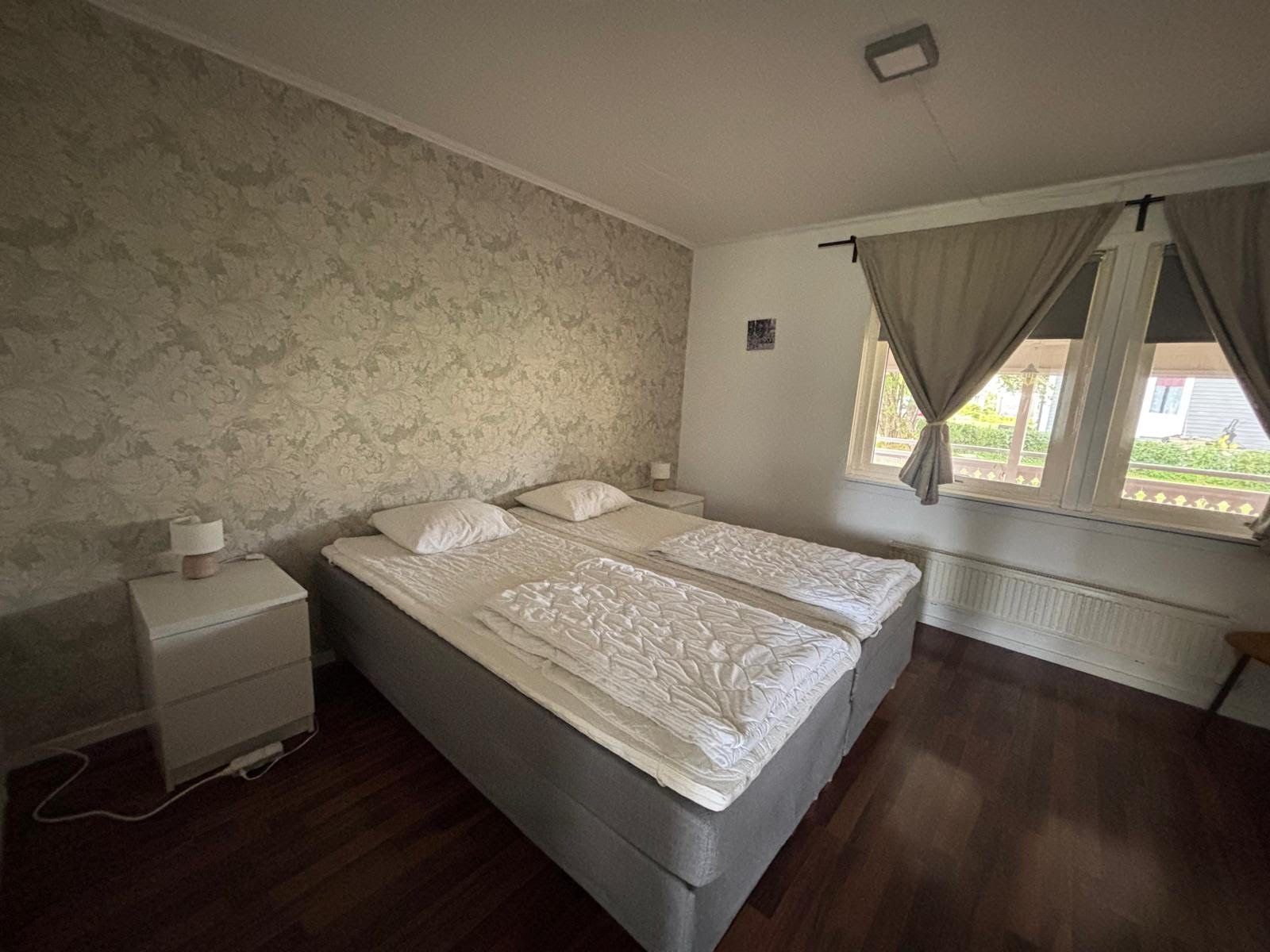 Une chambre à coucher avec un lit double, une table de chevet, une commode, une fenêtre avec des rideaux et un radiateur.
