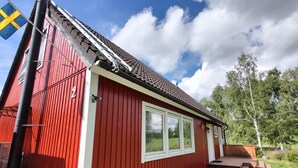 Un edificio in legno rosso con una finestra e una porta bianche, una bandiera e un vialetto sterrato.