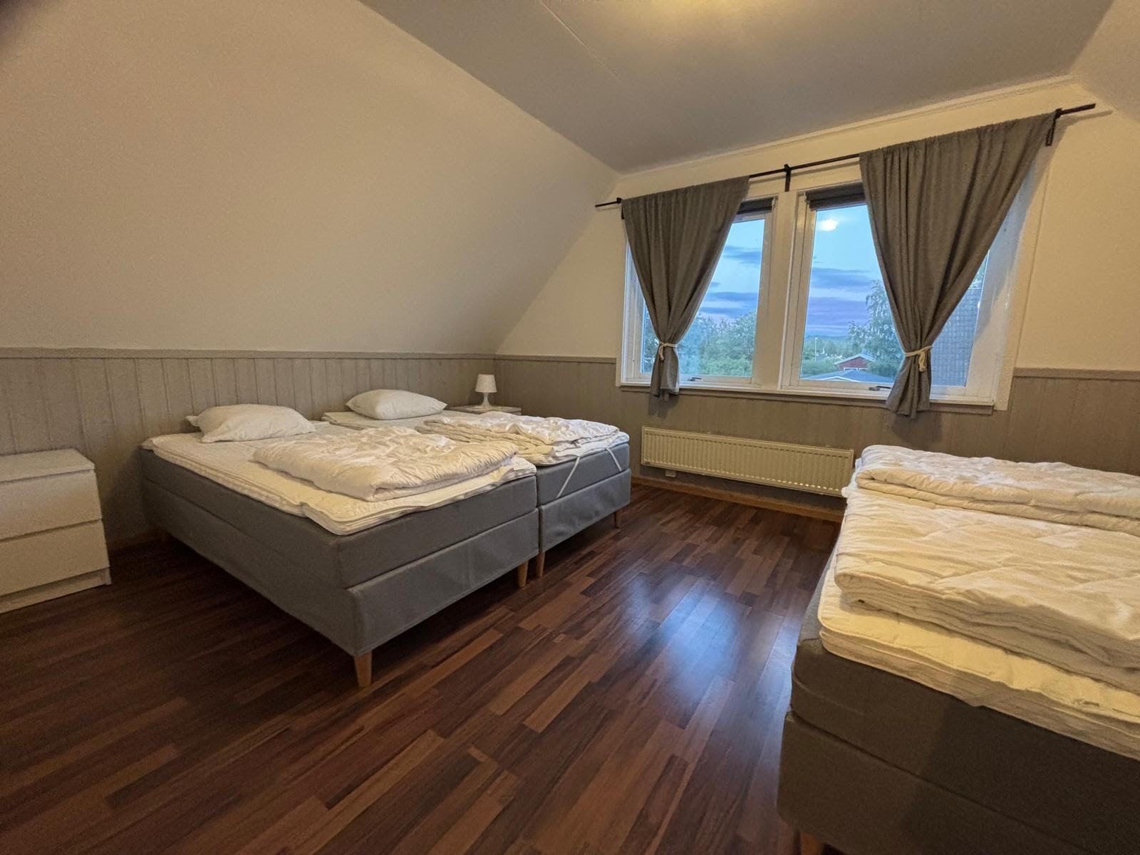 A bedroom with a single bed, a bedside table, a window with curtains, and a radiator.
