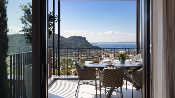A balcony with a table set for dining, overlooking a scenic view of a coastal town and mountains.