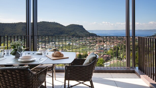 A balcony with a table set for dining, overlooking a scenic view of hills and a body of water. - Apartment Sole in Residence Villa Miralago (Costermano)