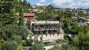 A modern building with a rooftop terrace, surrounded by lush greenery and residential houses in the background. - Apartment Sole in Residence Villa Miralago (Costermano)