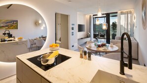 A modern kitchen with a marble countertop, a built-in stove, and a sink. There is a dining area with a round table and chairs, and a sofa in the background. - Apartment Sole in Residence Villa Miralago (Costermano)