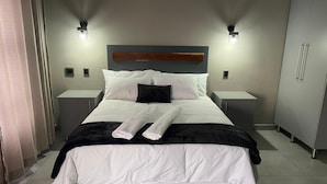 Ein ordentlich bezogenes Bett mit weißen Leinen und zwei gefalteten Handtüchern, ein dunkles Kopfteil, zwei Nachttische mit Lampen und ein Schrank zur Rechten.