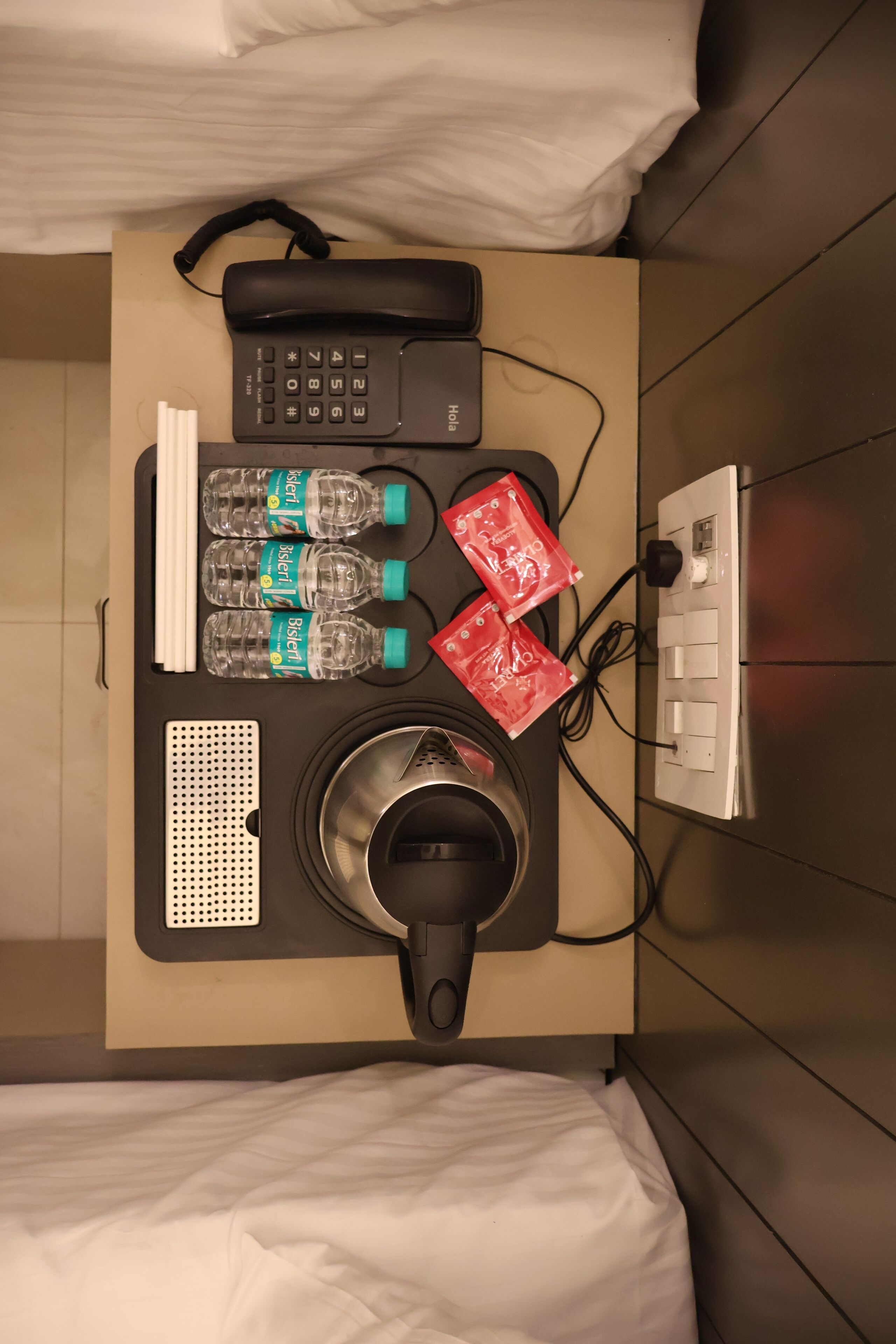 A hotel room with a bed, a desk with a phone, a kettle, a water bottle, a red packet, and a power outlet.