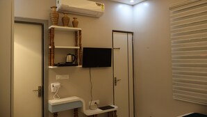 Ett hotellrum med en platt-TV som är monterad på väggen, ett litet sidobord med en telefon och en laddare, en hylla med dekorativa föremål och en luftkonditioneringsanläggning ovanför TV:n.