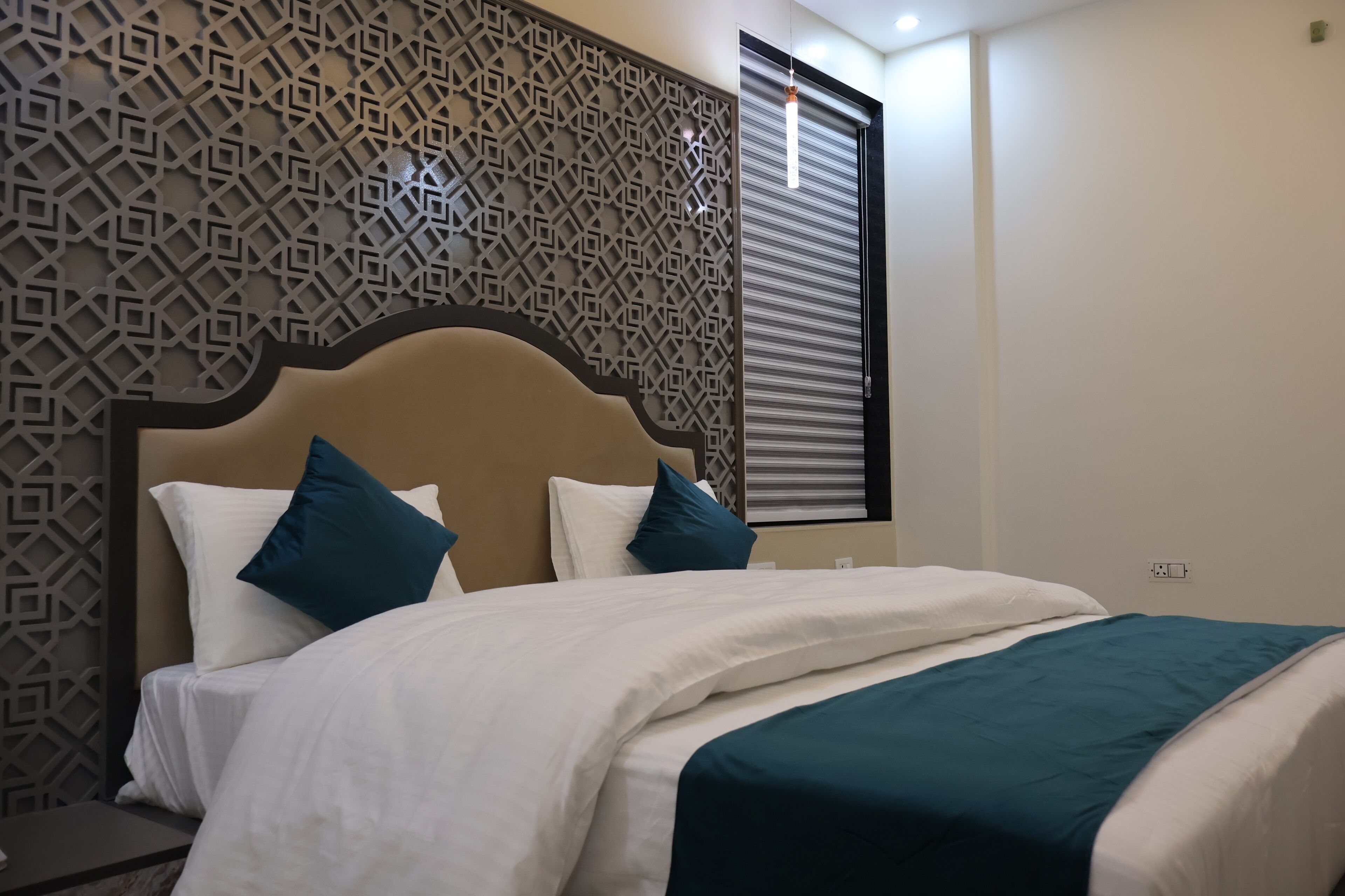 A neatly made bed with white and teal bedding, a decorative wall panel, and a window with blinds.