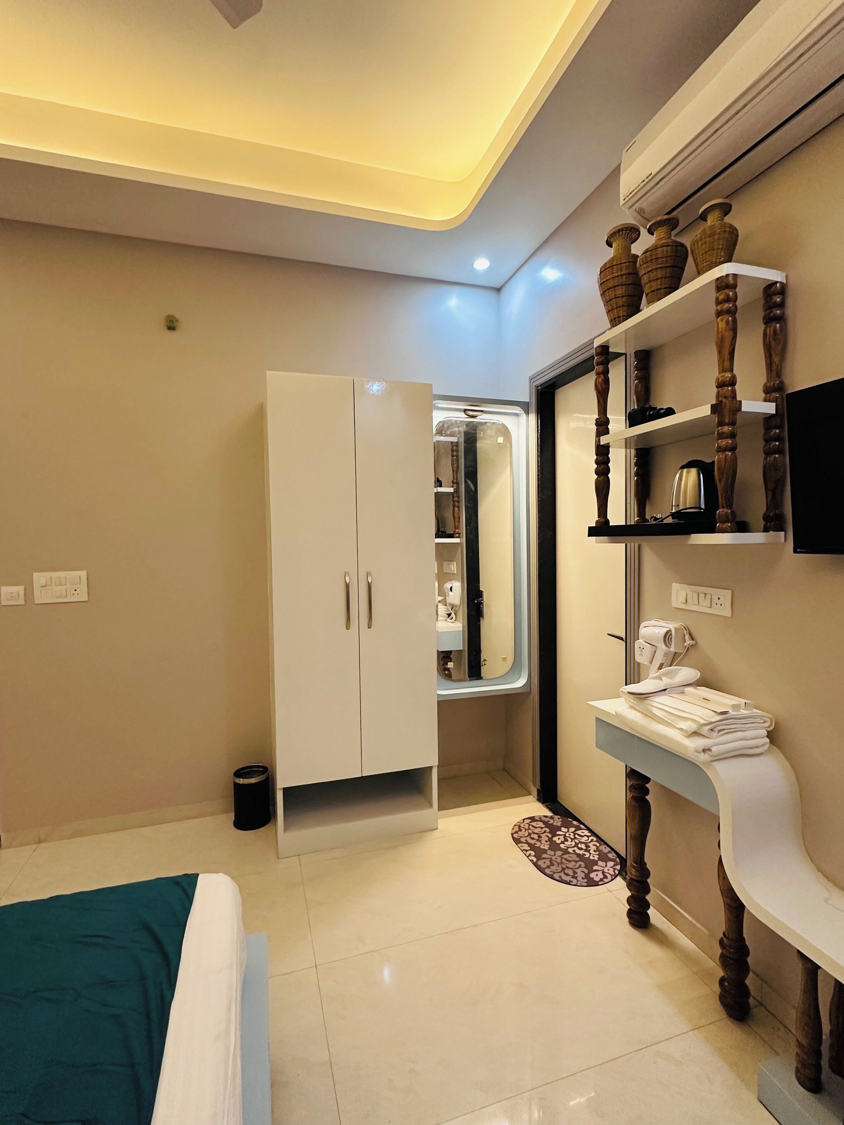 Quarto de hotel moderno com cama, guarda-roupa, penteadeira com espelho, prateleira com artigos de higiene pessoal e televisão na parede.