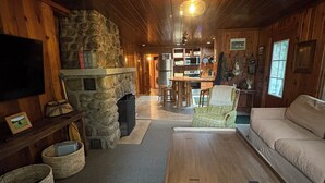 Interior acolhedor de cabana com lareira de pedra, móveis de madeira e televisão fixada na parede.