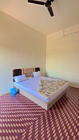 Una cama individual con una colcha estampada, un ventilador de techo y un cubo azul sobre un suelo tejido.