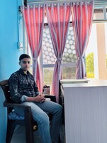 Una persona sentada en un escritorio con una ventana al fondo, cortinas cerradas y una pequeña mesa con una libreta.