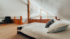 Ett sovrum med ett sluttande tak, en säng med en svartvit mönstrad kudde, en trästol och utsikt över naturen genom stora fönster.