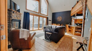 Ett mysigt vardagsrum med en öppen spis, en soffa, en matplats och ett stort fönster med utsikt över omgivningen.