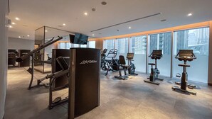 A modern gym with cardio equipment, a mirrored wall, and large windows offering a city view.