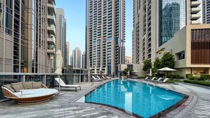 A swimming pool area with lounge chairs and a sunbed, surrounded by high-rise buildings.