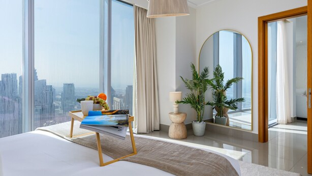 A modern bedroom with a large bed, a bedside table with a magazine and fruit, a potted plant, a mirror, and a view of the city through large windows.
