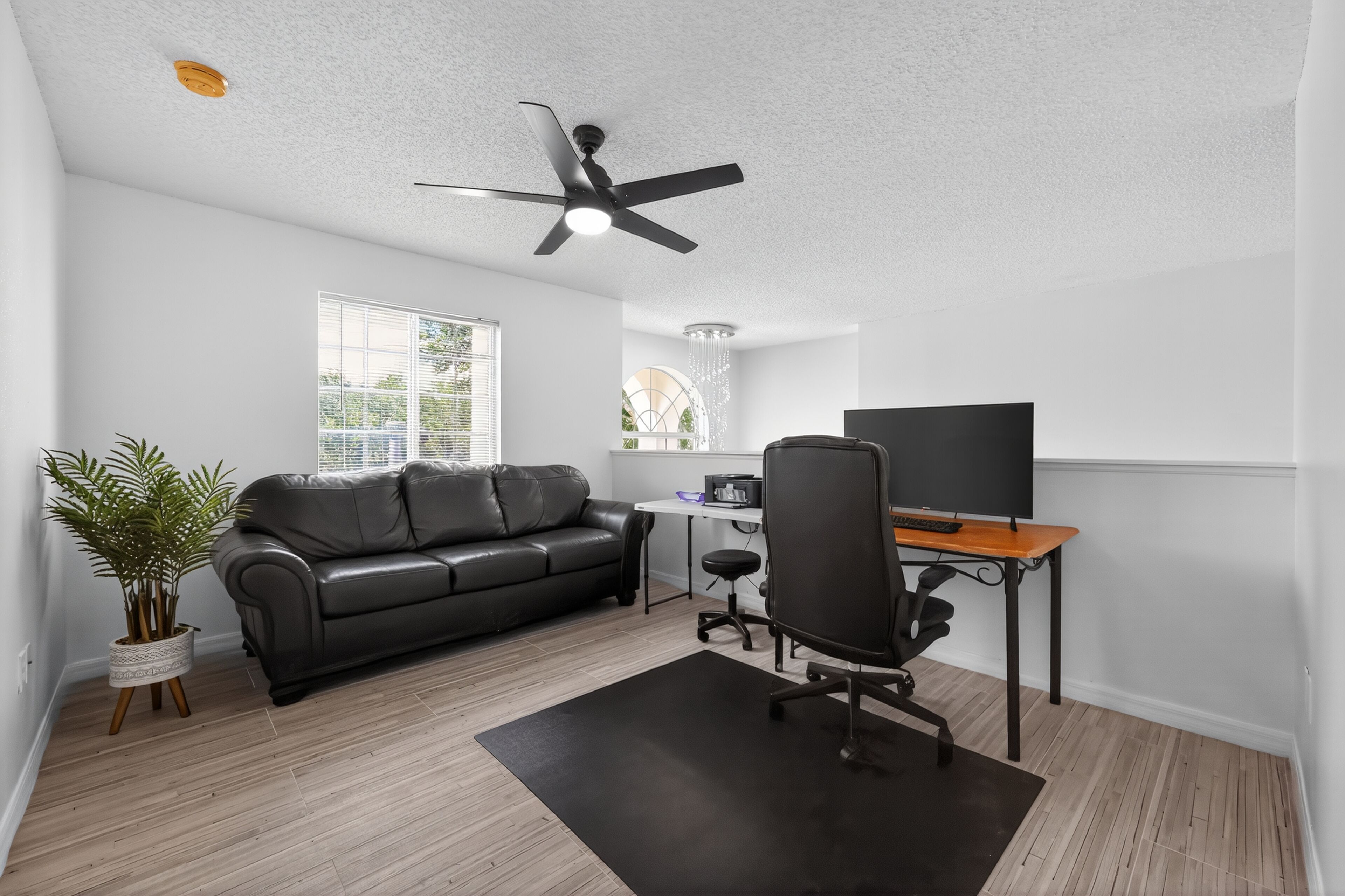 A modern living room with a black leather sofa, a wooden desk with a computer and monitor, a black office chair, a ceiling fan, and a potted plant.